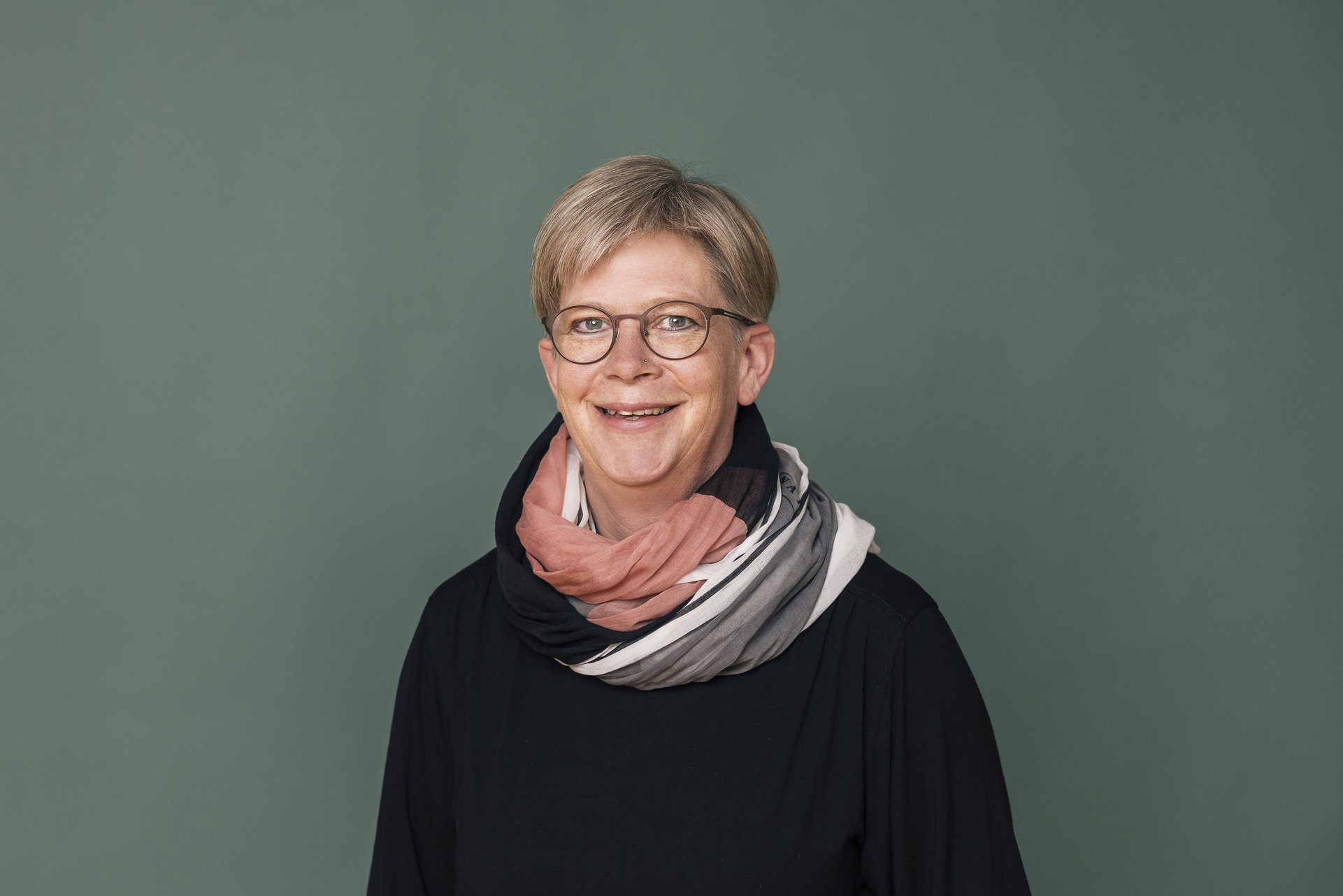 Eine Frau mit kurzen blonden Haaren. Sie trägt eine Brille und ein buntes Tuch. Sie lacht